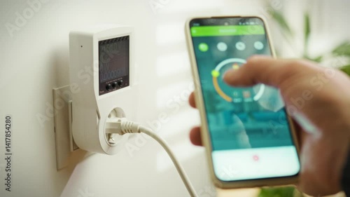 A person is using a smartphone to monitor energy usage via a smart plug device mounted on the wall. This activity occurs in a contemporary home, highlighting a focus on energy efficiency