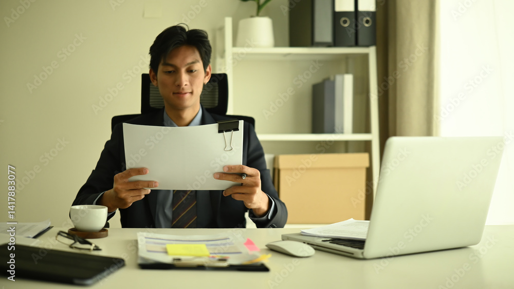 A professional businessman in a suit sits at a modern office desk, reviewing important documents. Business planning, data analysis, and corporate strategy in focus A professional businessman in a suit sits at a modern office desk, reviewing important documents. Business planning, data analysis, and corporate strategy in focus