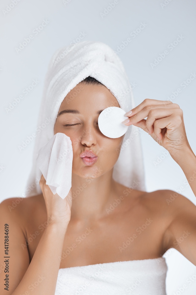 Close-up of a playful woman with a towel wrapped around her hair, making a kissy face while using a cotton pad and cleansing wipe during her skincare routine. Perfect for capturing fun and