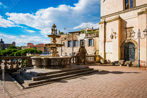 Ancient fountain standing in Piazza della Rocca, Viterbo, Italy, casting shadows on the brick pavement, with historic buildings and a glimpse of the cityscape under a bright sky