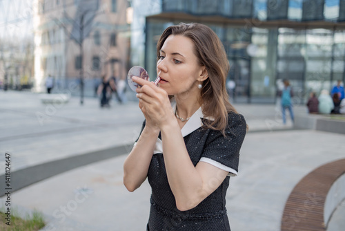 A young woman applying lipstick in a public place, wearing a black dress with white collar. She is focused on her reflection in a compact mirror. The background shows a modern cityscape with people 