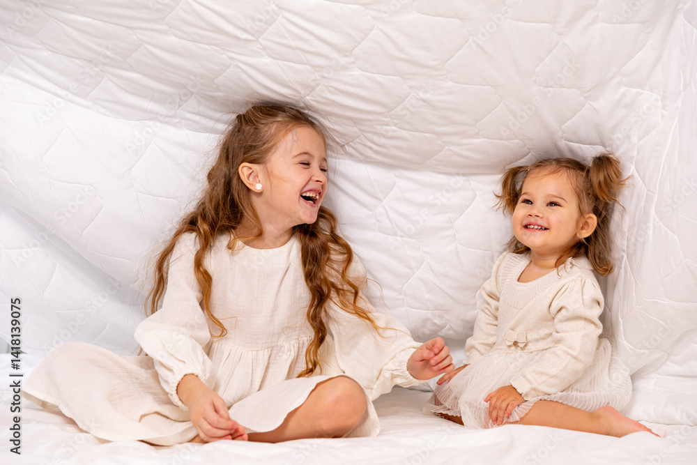 Children sisters in bed under the blanket having fun, smiling. Happy little girl and her newborn baby sister hiding under the white blanket. Childhood concept. Copy space. High quality photo