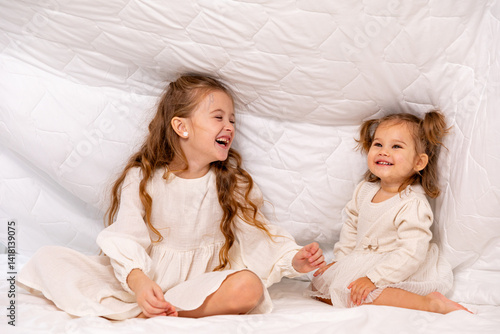 Obraz na płótnie Children sisters in bed under the blanket having fun, smiling. Happy little girl and her newborn baby sister hiding under the white blanket. Childhood concept. Copy space. High quality photo w salonie