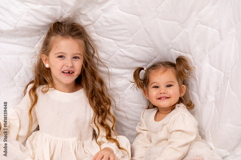Children sisters in bed under the blanket having fun, smiling. Happy little girl and her newborn baby sister hiding under the white blanket. Childhood concept. Copy space. High quality photo