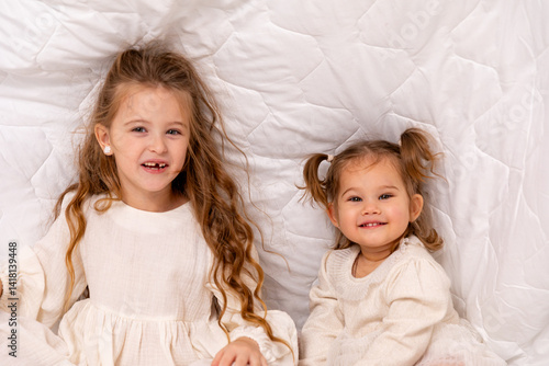 Children sisters in bed under the blanket having fun, smiling. Happy little girl and her newborn baby sister hiding under the white blanket. Childhood concept. Copy space. High quality photo