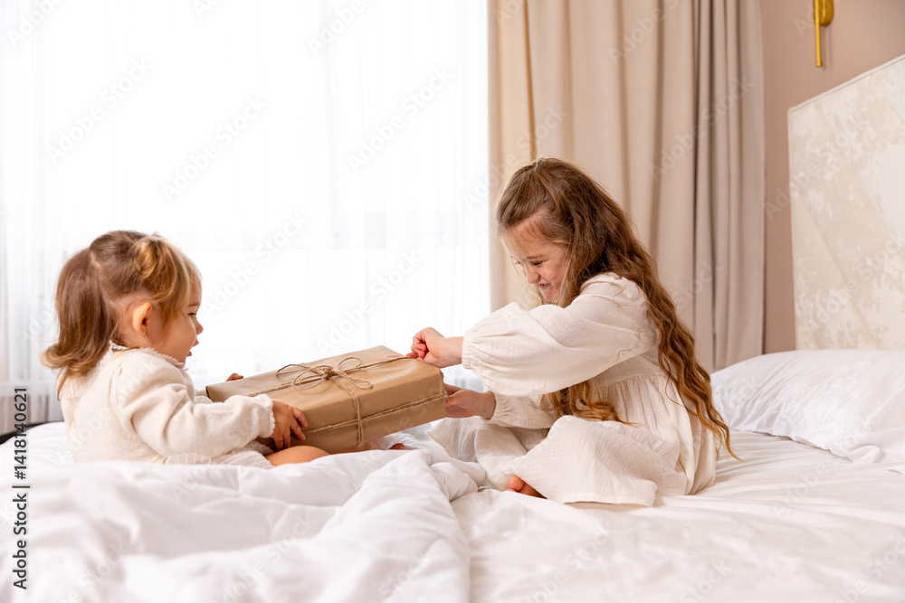 Two sisters wearing white dresses, sitting on a bed with white bedding, pulling big wrapped gift box, fighting, cannot share a present on the holiday morning at home