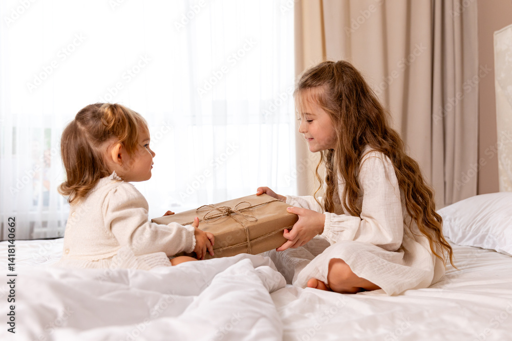 Two sisters wearing white dresses, sitting on a bed with white bedding, pulling big wrapped gift box, fighting, cannot share a present on the holiday morning at home