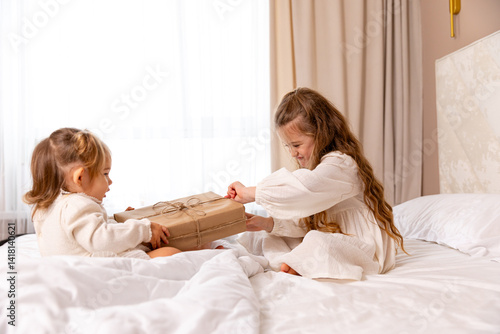 Obraz na płótnie Two sisters wearing white dresses, sitting on a bed with white bedding, pulling big wrapped gift box, fighting, cannot share a present on the holiday morning at home w salonie