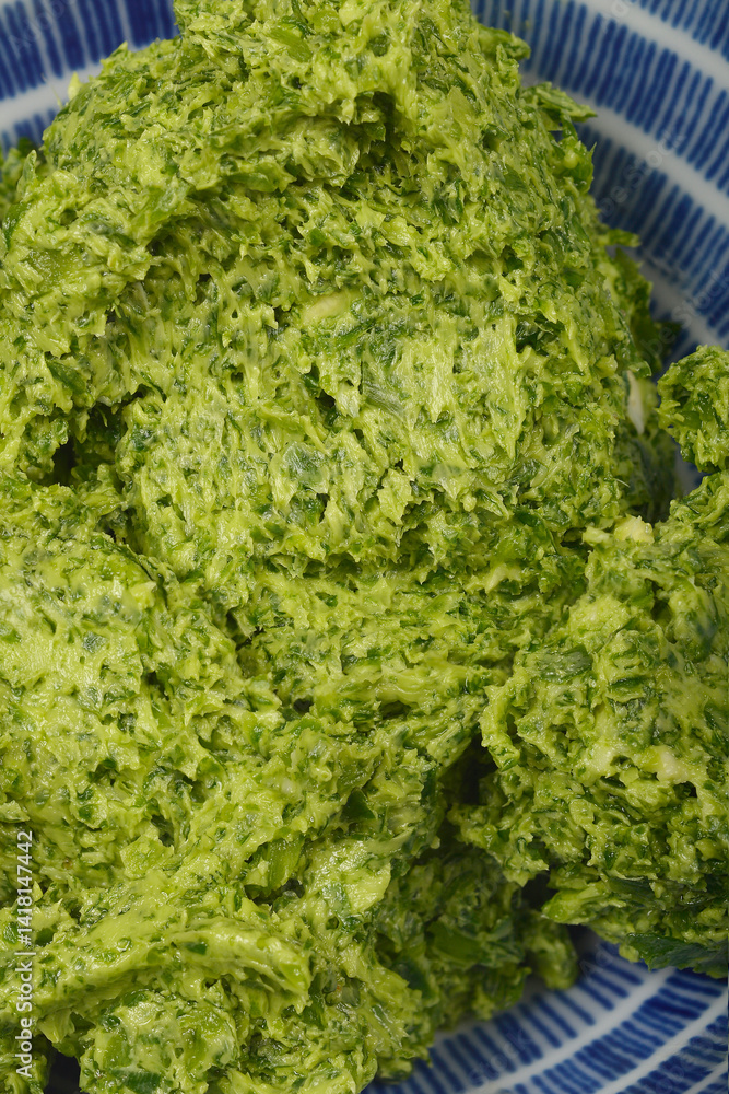 A close-up, top-down shot reveals the textured surface of vibrant green wild garlic (Allium ursinum)) butter within a blue and white striped bowl