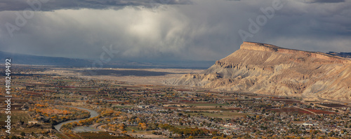 The Grand Valley, the Colorado River, Mt. Garfield, and the Book Cliffs are viewed from a cliff high above Palisade, Colorado, as storms lurk beyond.