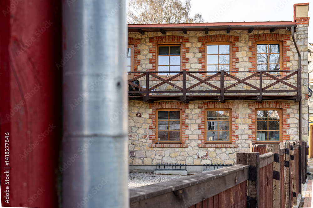 Obraz premium Stone and Brick House with Wooden Balcony Framed by Metal and Wooden Posts – Urban Architecture Perspective in Soft Natural Light