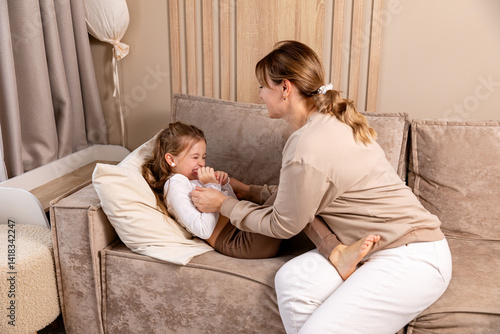 Obraz na płótnie Happy mom tickling kid daughter, laughing together on couch. Cheerful mother playing funny game with little cute girl at home, having fun, relaxing. Happy family. High quality photo w salonie