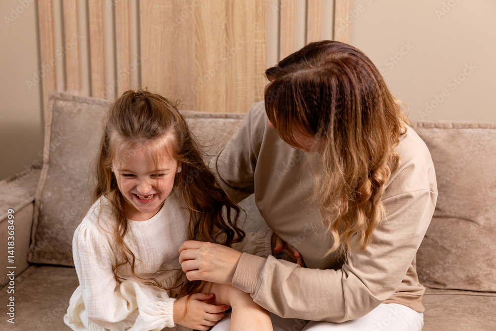 Happy mom tickling kid daughter, laughing together on couch. Cheerful mother playing funny game with little cute girl at home, having fun, relaxing. Happy family. High quality photo