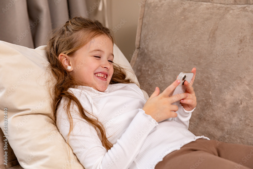 Smiling little child lying on soft couch at living room, look at phone screen use game app contact mates at social network. Funny small girl engaged in watching online channel for children. Copy space