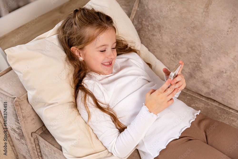 Smiling little child lying on soft couch at living room, look at phone screen use game app contact mates at social network. Funny small girl engaged in watching online channel for children. Copy space