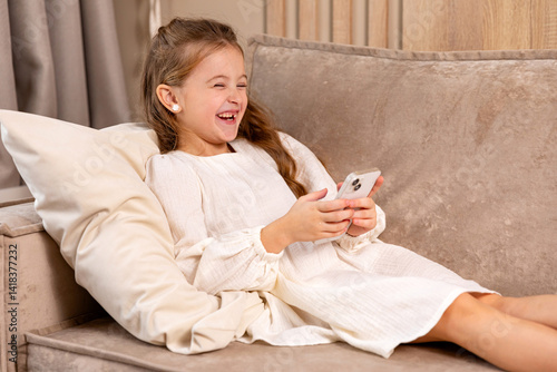 Obraz na płótnie Laughing little child lying on soft couch at living room, look at phone screen use game app contact mates at social network. Small girl engaged in watching online channel for children. Copy space w salonie
