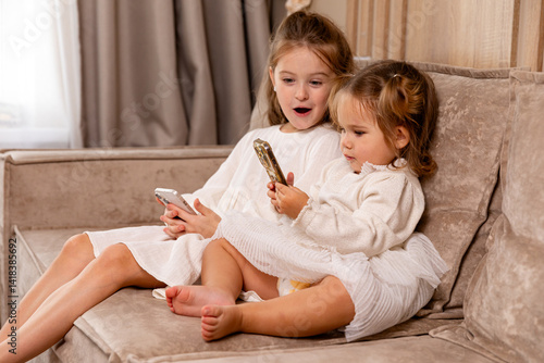 Obraz na płótnie Two little girls sisters sit on soft couch at living room, hold smartphones, use game app and social network. Kids engaged in watching online channel for children, cartoons. High quality photo w salonie