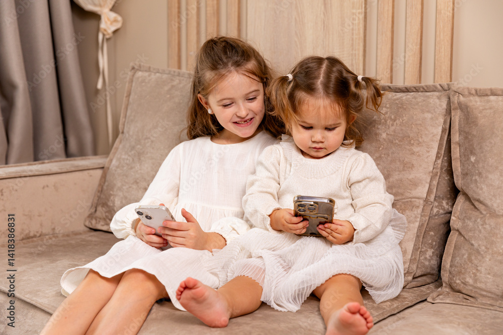 Two little girls sisters sit on soft couch at living room, hold smartphones, use game app and social network. Kids engaged in watching online channel for children, cartoons. High quality photo