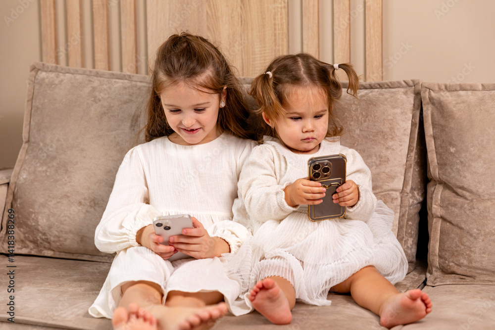 Two little girls sisters sit on soft couch at living room, hold smartphones, use game app and social network. Kids engaged in watching online channel for children, cartoons. High quality photo