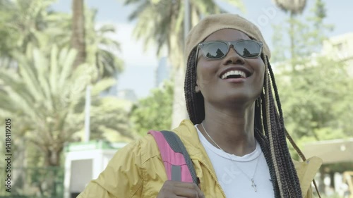 Beautiful Black Latina woman with braids, wearing a yellow jacket, beret, and sunglasses, smiling with curiosity while sightseeing in a Chilean city, carrying a backpack and enjoying the urban vibe