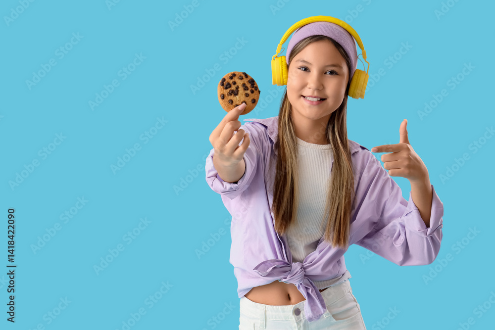 Cute teenage girl in headphones pointing at delicious cookie on blue background