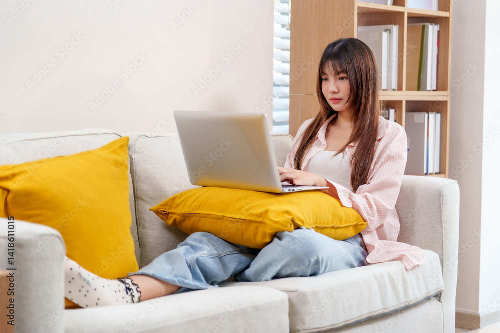 young woman is comfortably working from home while sitting on cozy sofa with laptop on her lap. She appears focused and relaxed, surrounded by bright and inviting living space.freeelancer young woman is comfortably working from home while sitting on cozy sofa with laptop on her lap. She appears focused and relaxed, surrounded by bright and inviting living space.freeelancer
