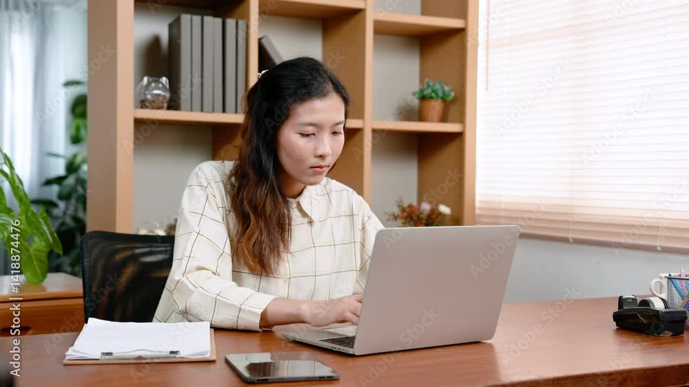 Asian woman sitting at desk, room in office, there was laptop computer on table in front, using hands write various documents on paper, turned looked at computer screen with questioning look on face. Asian woman sitting at desk, room in office, there was laptop computer on table in front, using hands write various documents on paper, turned looked at computer screen with questioning look on face.