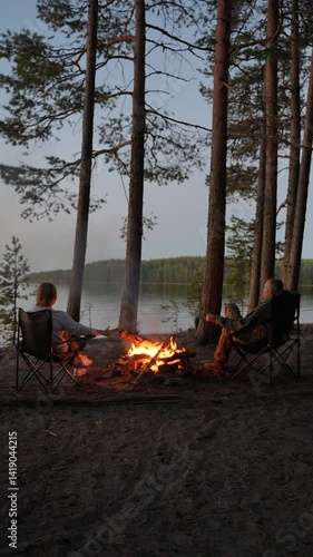Tourists are warming themselves by the campfire with a view of the lake. A married couple is relaxing on the shore of a forest lake by the fire. A romantic evening in nature.