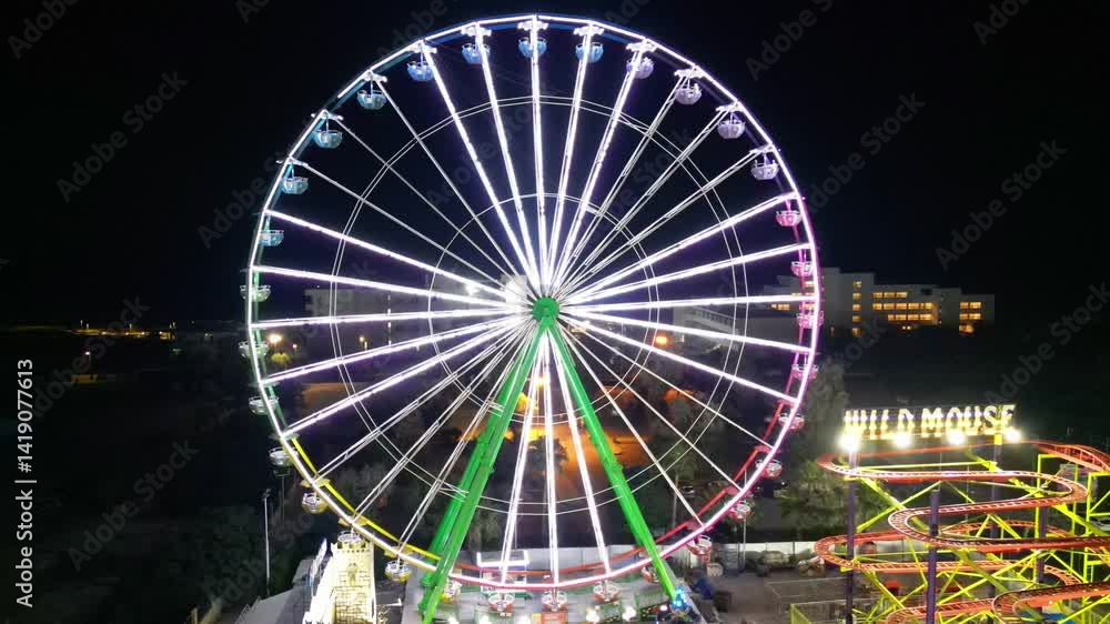 Large Ferris wheel glowing with colorful lights beside Wild Mouse ride at night in Ayia Napa. Ayia Napa, Cyprus Large Ferris wheel glowing with colorful lights beside Wild Mouse ride at night in Ayia Napa. Ayia Napa, Cyprus