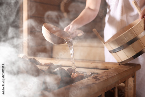 Fotografija Woman pouring water onto stones surrounded by steam in sauna, closeup
