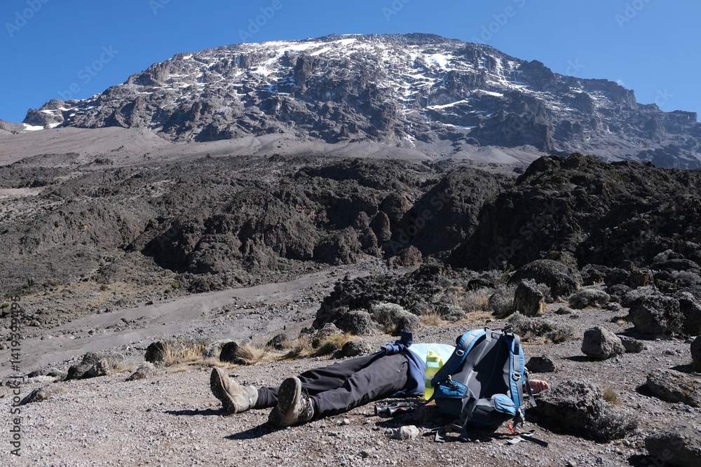 Silhouette of lying down exhausted man next ot backpack and Kilimanjaro peak in background, on trail from Barranco Hut to Barafu Camp, during trekking to Kilimanjaro Peak, Machame Route. Tanzania