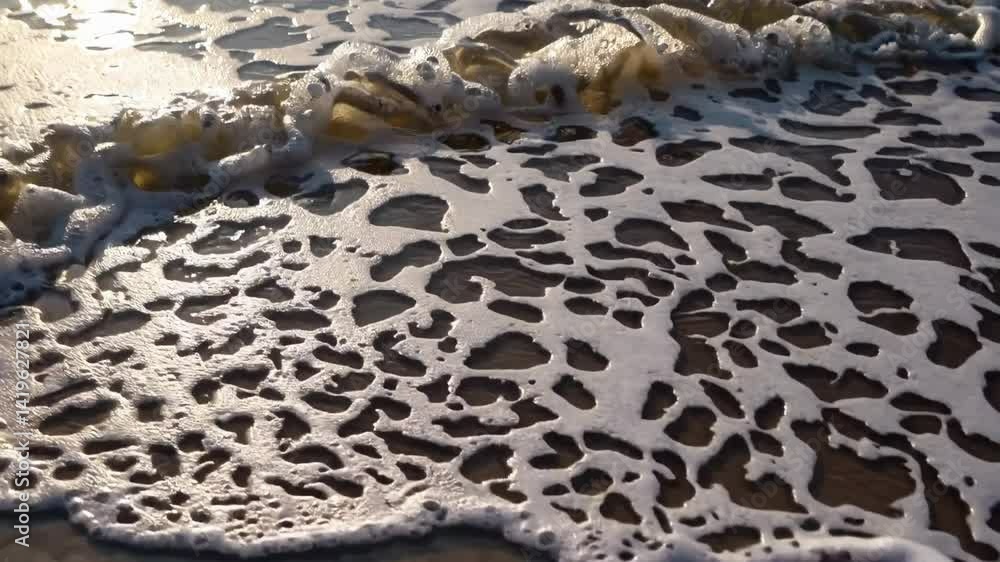 Sunlight glints off the wet sand as delicate waves create intricate patterns of foam, receding gently and leaving behind a mesmerizing, textured surface