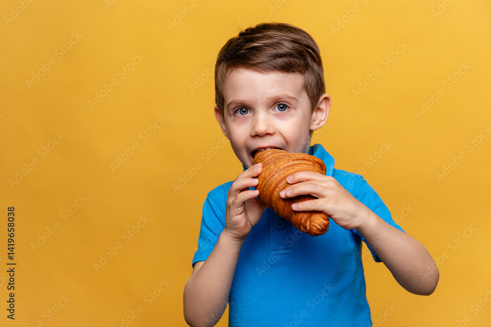 Happy smiling little boy in blue t-shirt eating French croissant on yellow background. Copy space. High quality photo. French breakfast concept.