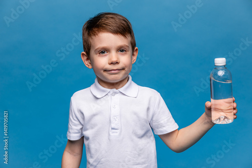 Obraz na płótnie A happy little boy drinks water for health on a blue background. The child is holding a water bottle. A kid with a plastic bottle. Children's activity and energy, generation Z. body hydration concept w salonie