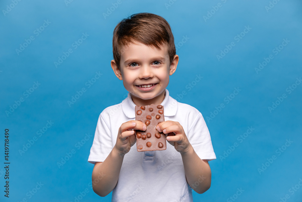 Cute little boy, 3-4 years old, wearing white t-shirt, eating chocolate bars with nuts on blue background, closeup. The concept of children's happiness and love for sweets. High quality photo