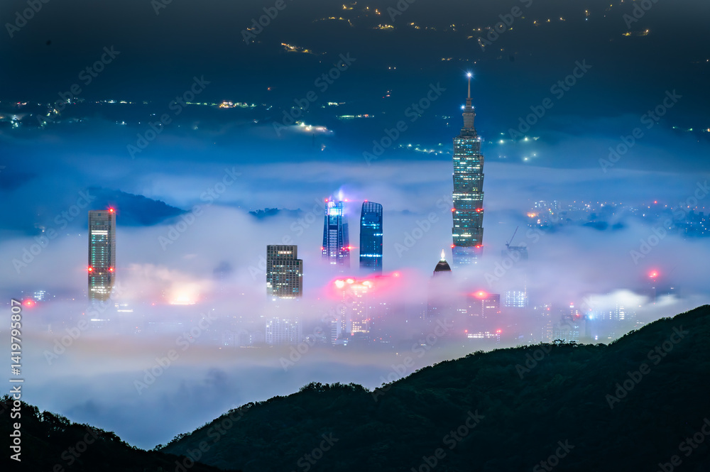 Obraz premium Night view from Datun Mountain, Taipei, Taiwan during springtime, showcasing the city skyline with prominent skyscrapers partially covered by low-lying mist. City lights pierce through the fog.