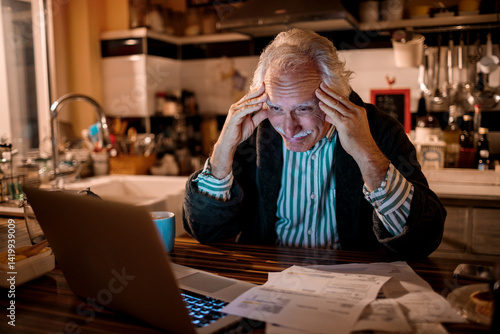 Senior man stressed while looking at bills on laptop at night in kitchen