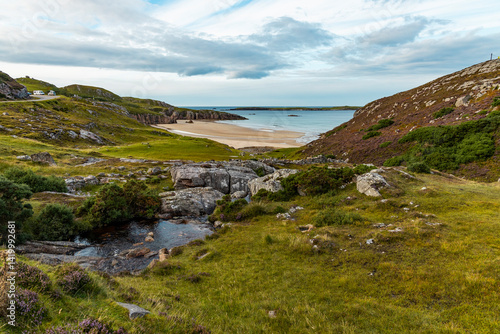 Ceannabeinne Beach shines under a clear summer sky. Crystal-clear waters, golden sand with a heart drawn on it, and lush green grass create a pristine and romantic coastal escape.

