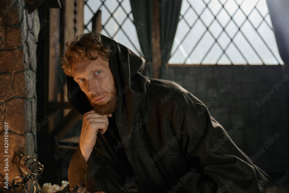 Portrait of serious pensive medieval monk in hooded robe sitting at table
