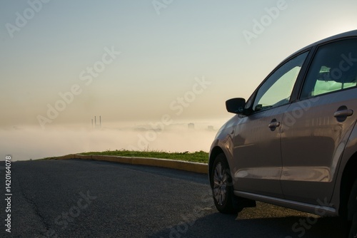 A clean, new car is parked on the side of the road on a mountain against the backdrop of a city in the fog