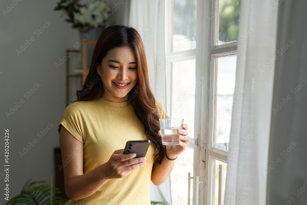 Connection and Technology. A woman checks her phone while enjoying a glass of water by the window. Connection and Technology. A woman checks her phone while enjoying a glass of water by the window.