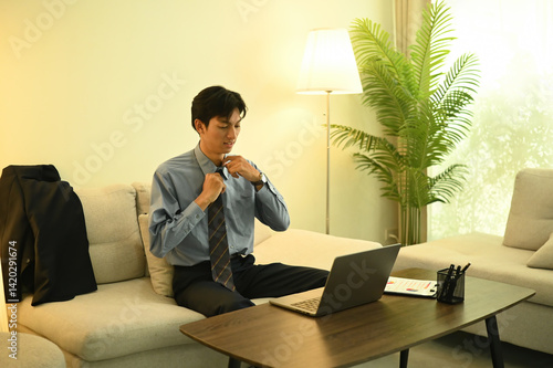 A young businessman arrives home, loosens his tie, and prepares to continue working on his laptop. A visual of dedication, stress, and the non-stop pace of modern working life