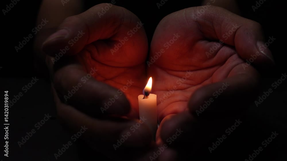 A person protecting the flame of the lit candle isolated on the black background. The candle flame illuminates the palms, symbolizing the bitterness of loss, death, and grief. A person protecting the flame of the lit candle isolated on the black background. The candle flame illuminates the palms, symbolizing the bitterness of loss, death, and grief.
