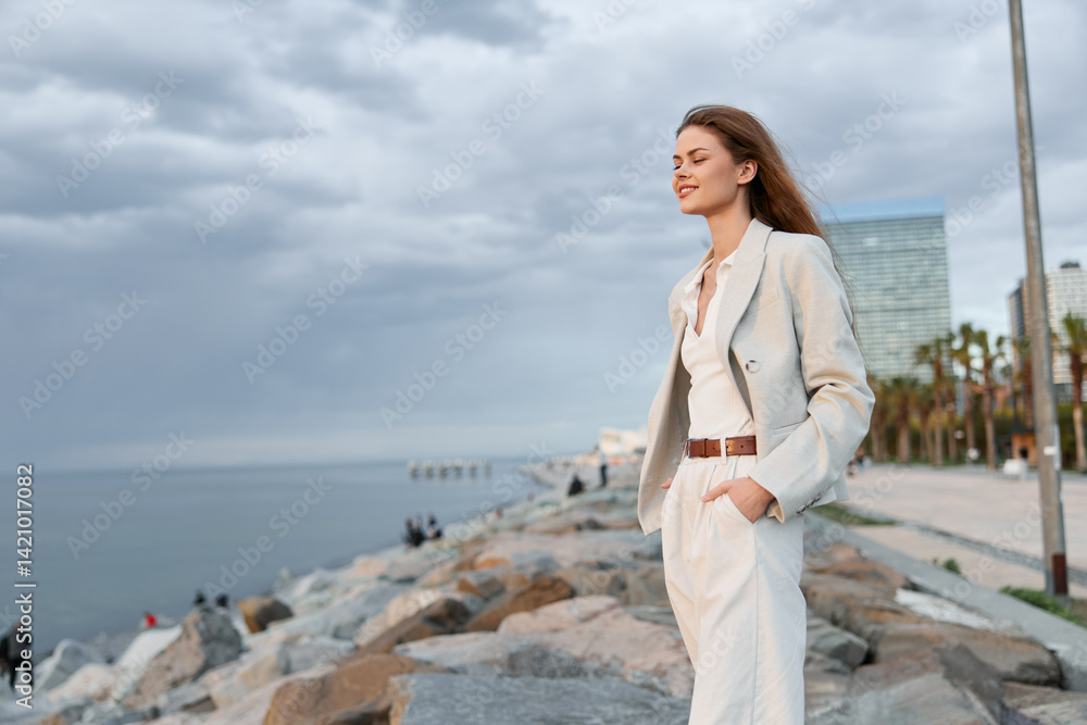 Young woman in beige pantsuit enjoying calm seaside breeze with modern city and cloudy sky background, fashion style, confident smiling woman outdoors Young woman in beige pantsuit enjoying calm seaside breeze with modern city and cloudy sky background, fashion style, confident smiling woman outdoors