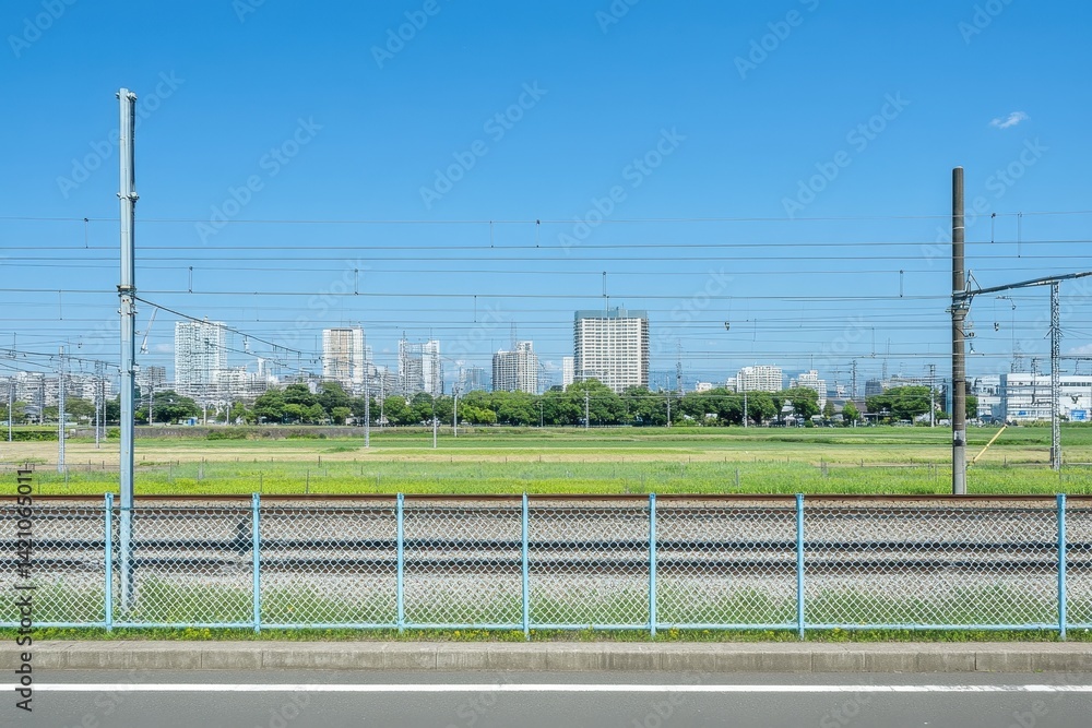 Fototapeta premium Urban landscape, rail tracks and open space. A view of a city skyline behind a grassy field and metal fence, with train tracks in the foreground. Clear, blue sky