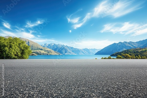 Fototapeta Naklejka Na Ścianę i Meble -  Empty asphalt road leading to a scenic mountain lake vista. Lush green foliage lines the shores of a tranquil lake, nestled amidst towering mountains under a vibrant blue sky