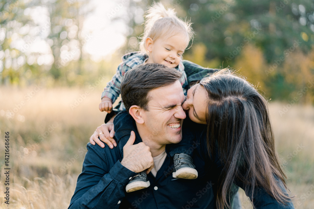 Obraz premium Mom hugs and kisses dad on the cheek from behind with a little girl on his shoulders