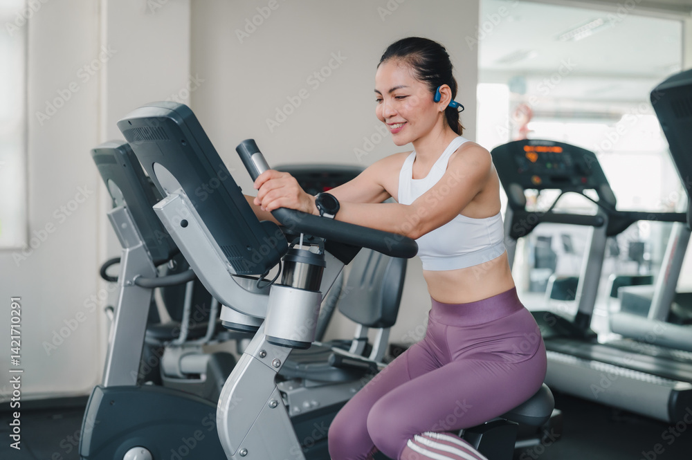 Fototapeta premium Empowered Woman Crushing Her Exercise Goals Smiling In Gym On An Exercise Bike, Toned Physique While Wearing White Sports Bra, Focused Workout With Fitness Equipment Smartwatch, Boosting Weight Loss