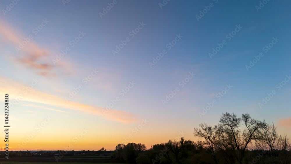 Obraz premium A tranquil sunset view over a rural landscape, showcasing vibrant colors in the sky at dusk, with silhouettes of trees framing the scene, ideal for nature-inspired themes.