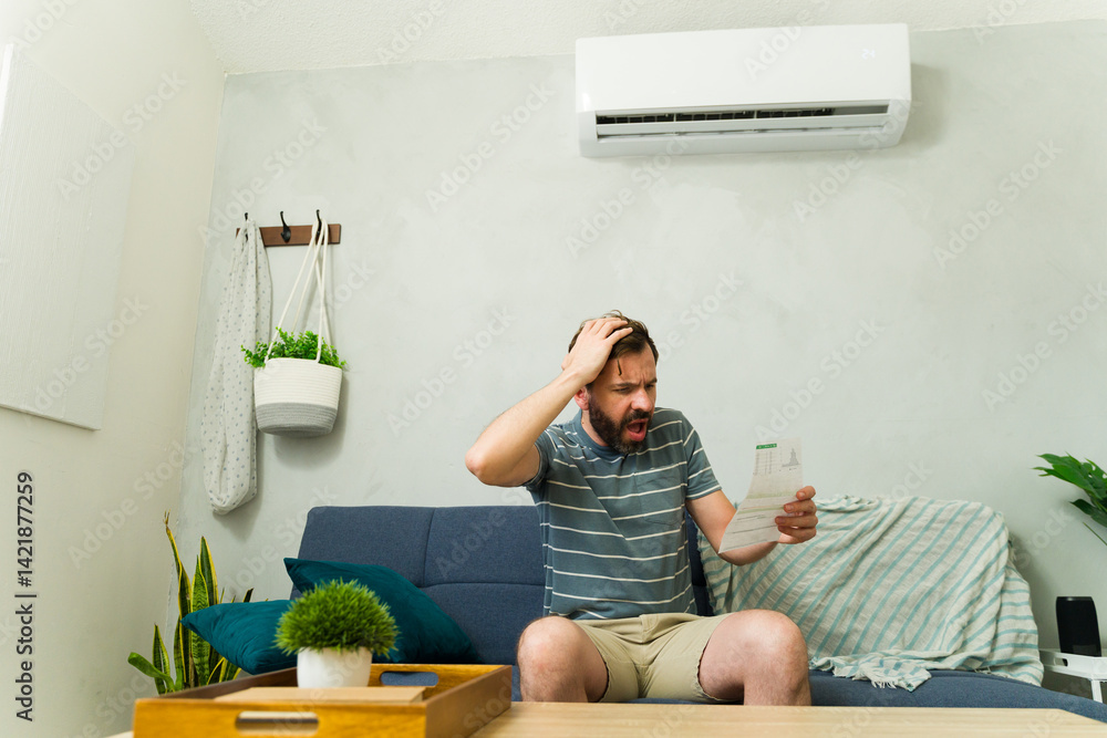Worried man holding electricity bill sitting on sofa at home with air conditioning unit